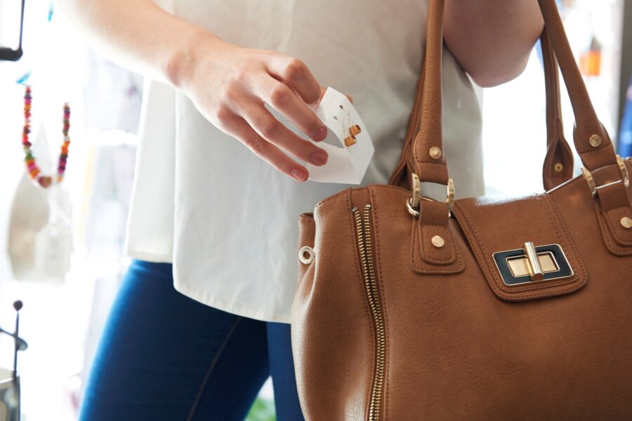 A woman in blue jeans and a white blouse steals an expensive pair of earrings, slipping them into a brown bag.